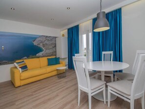 Ein Zimmer mit einem gelben Sofa, weißen Stühlen, einem runden Tisch und einem großen Fenster mit blauen Vorhängen.