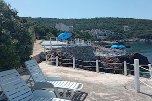 Eine Küstenlandschaft mit einer Steinmauer, Sitzgelegenheiten und Blick auf das Meer und die Hügel.