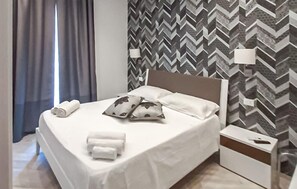 A neatly made bed with pillows and a white blanket, a bedside table with a lamp, and a patterned wall. - Lovely apartment in Donnalucata with WiFi (Donnalucata)