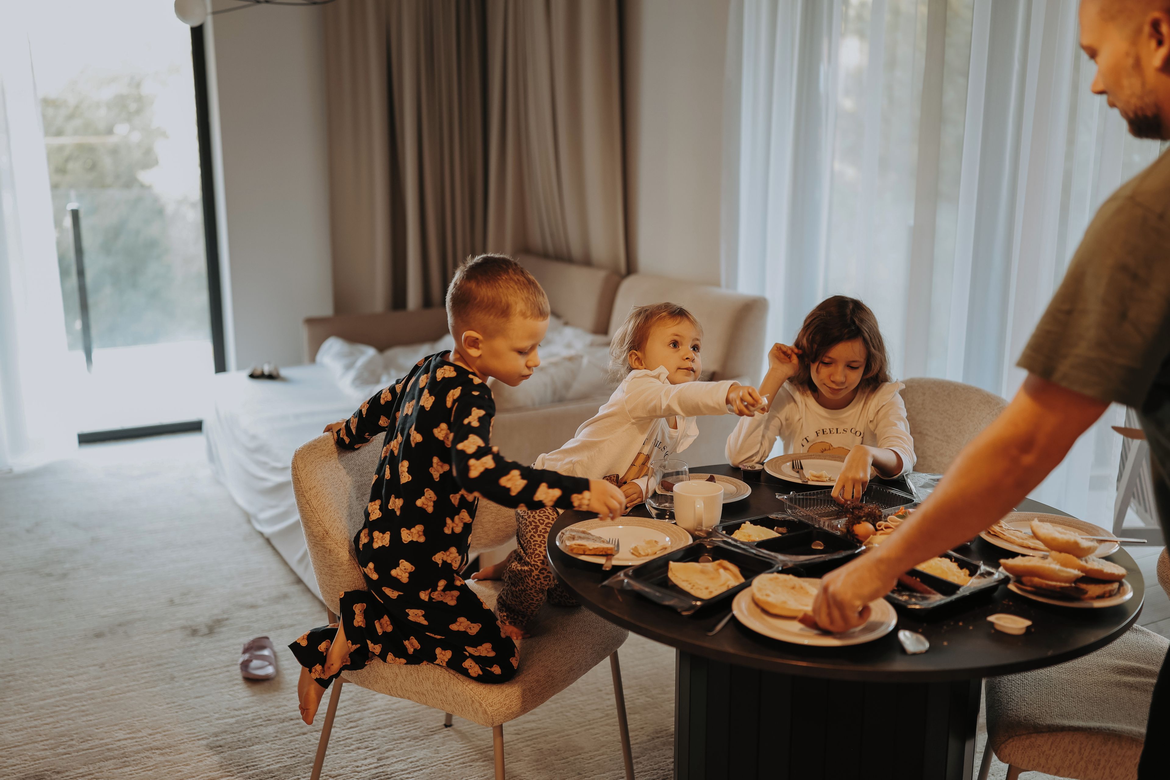 Une famille est assise autour d’une table ronde dans une chambre d’hôtel, avec un lit en arrière-plan.