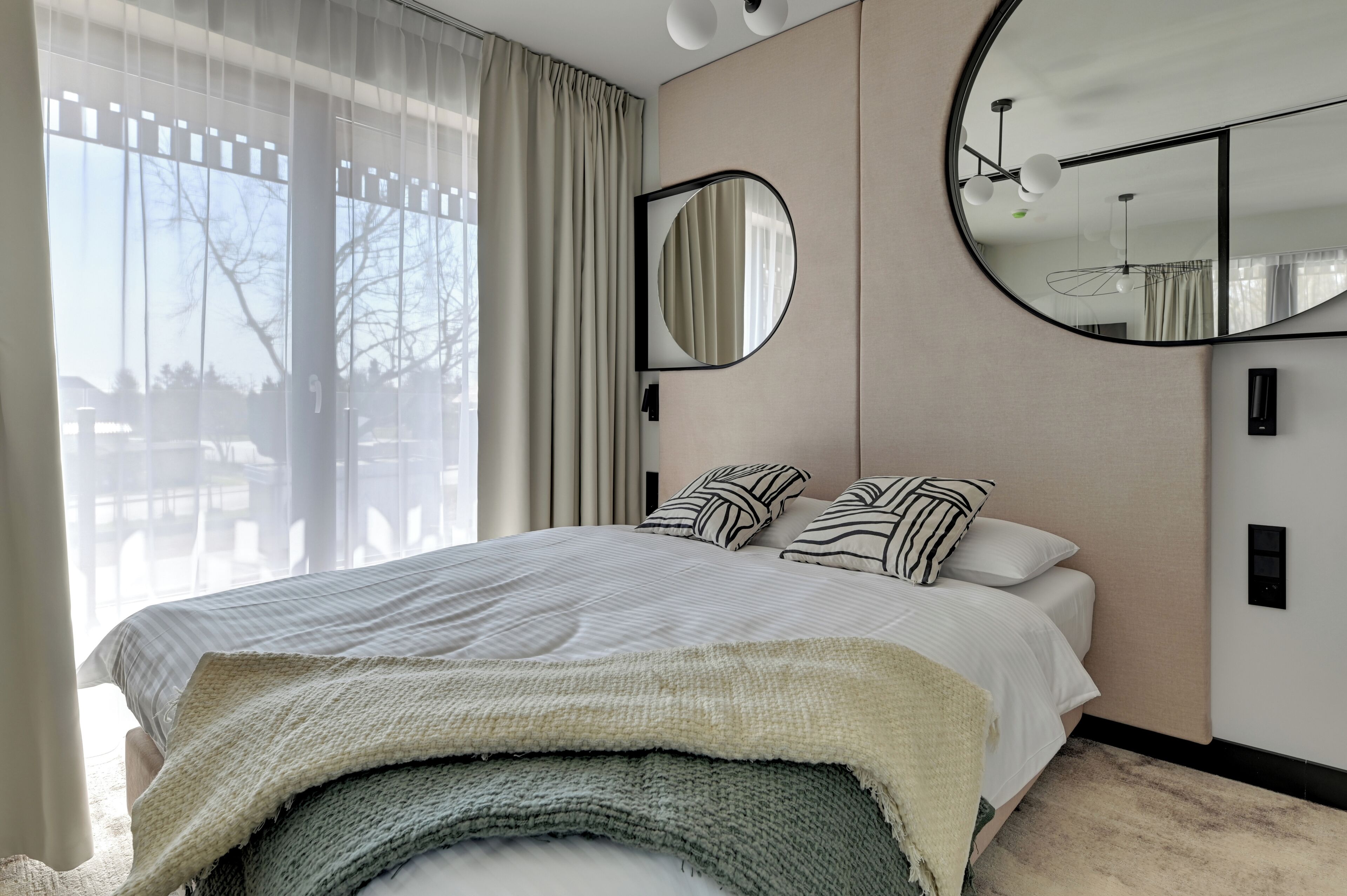 Ein modernes Schlafzimmer mit einem großen Bett, einem runden Spiegel und Blick ins Freie durch transparente Vorhänge.