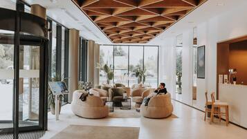 Un moderno vestíbulo de hotel con una zona de estar central, ventanas amplias y un diseño de techo llamativo.