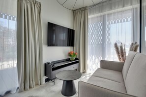 A modern living room with a sofa, a coffee table, a flat-screen TV mounted on the wall, a console table with a vase of flowers, and sheer curtains. - Grano Aparthotel Ustronie Morskie (Ustronie Morskie)