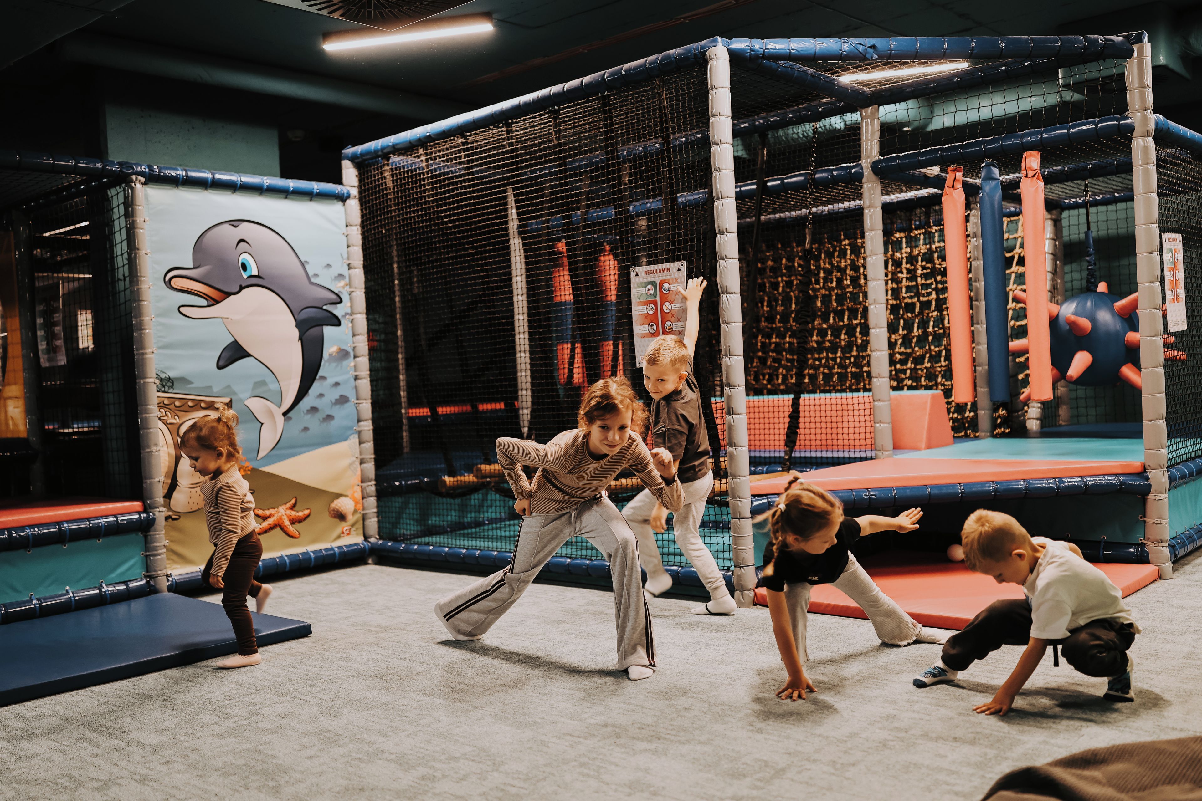 Zona de juegos infantiles con un mural de tiburón, estructuras acolchadas y una pared de escalar con red.