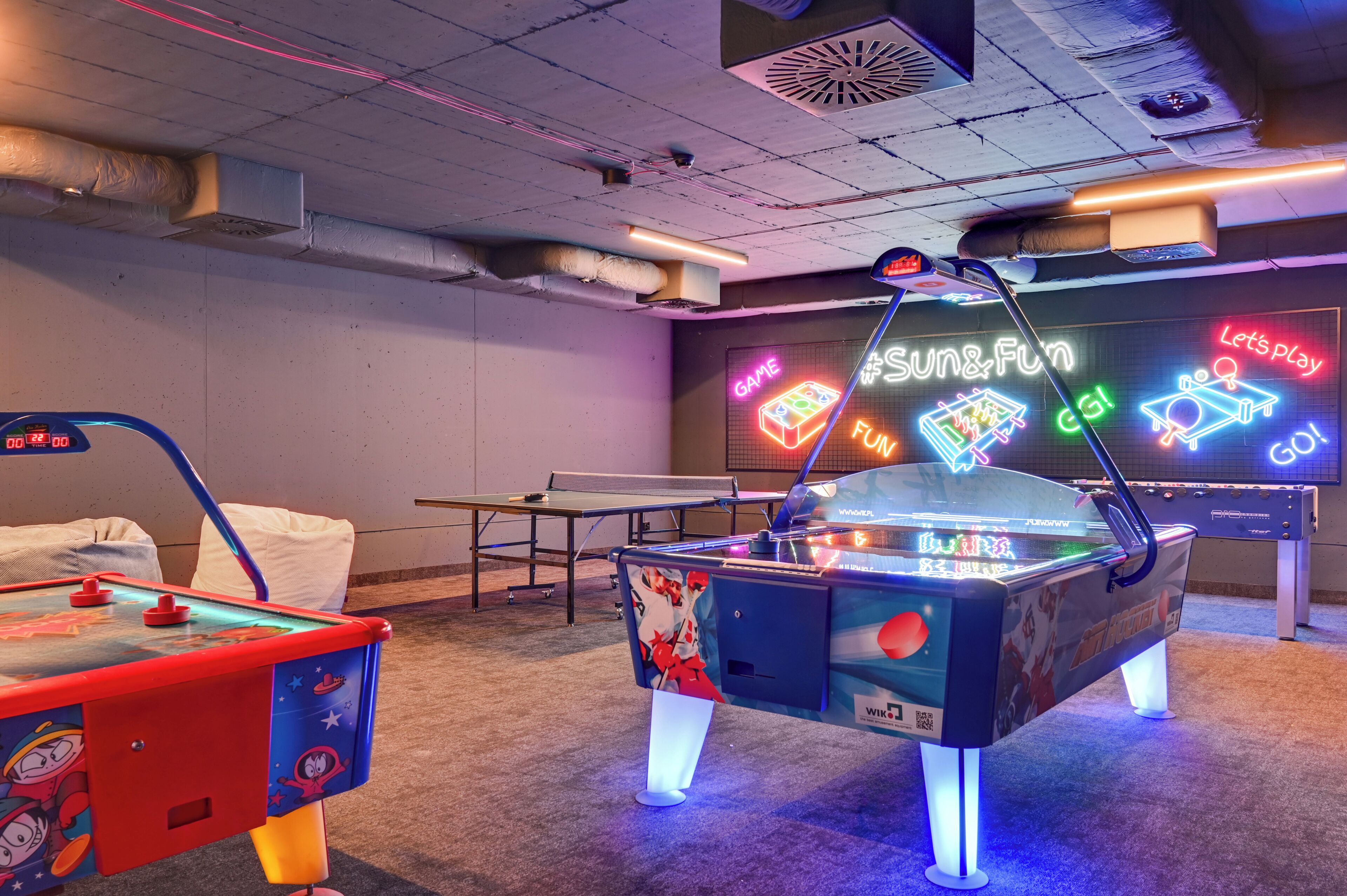 A room with ping pong tables, a foosball table, and a neon sign.