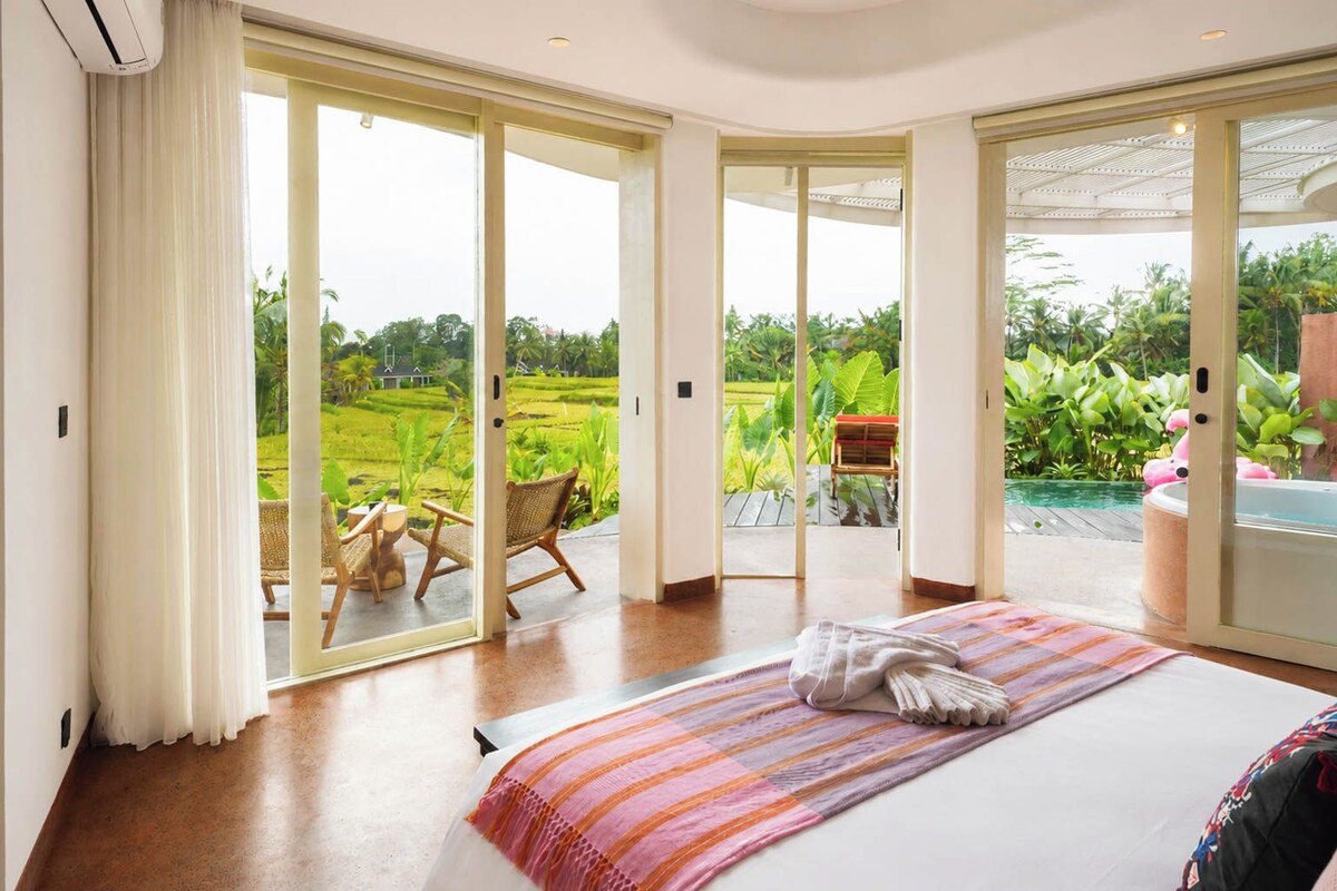 A bedroom with a large bed, a view of a lush garden, and a hot tub outside.