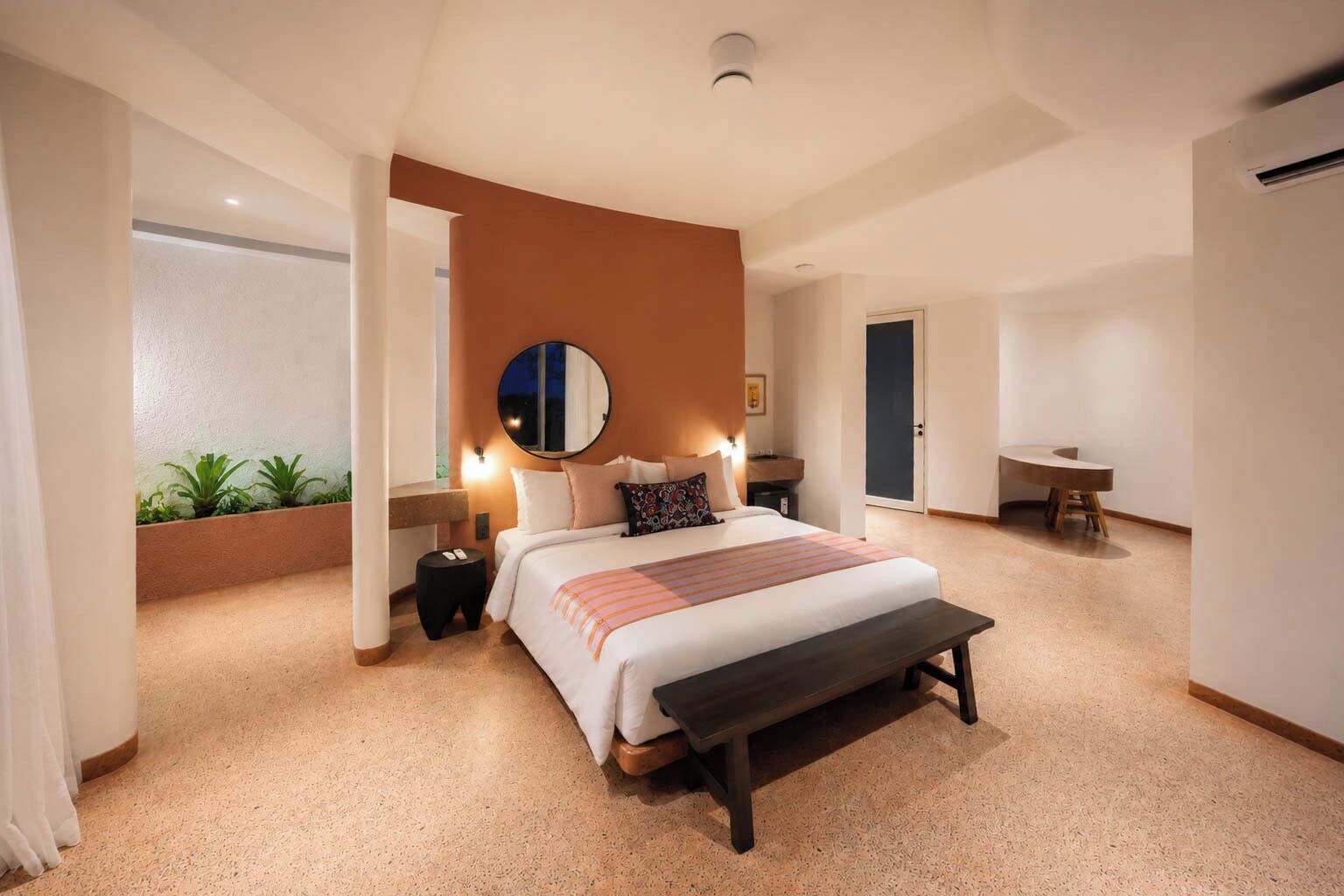 Un dormitorio con una cama grande, mesitas de noche, un banco, un espejo redondo y vistas al exterior.