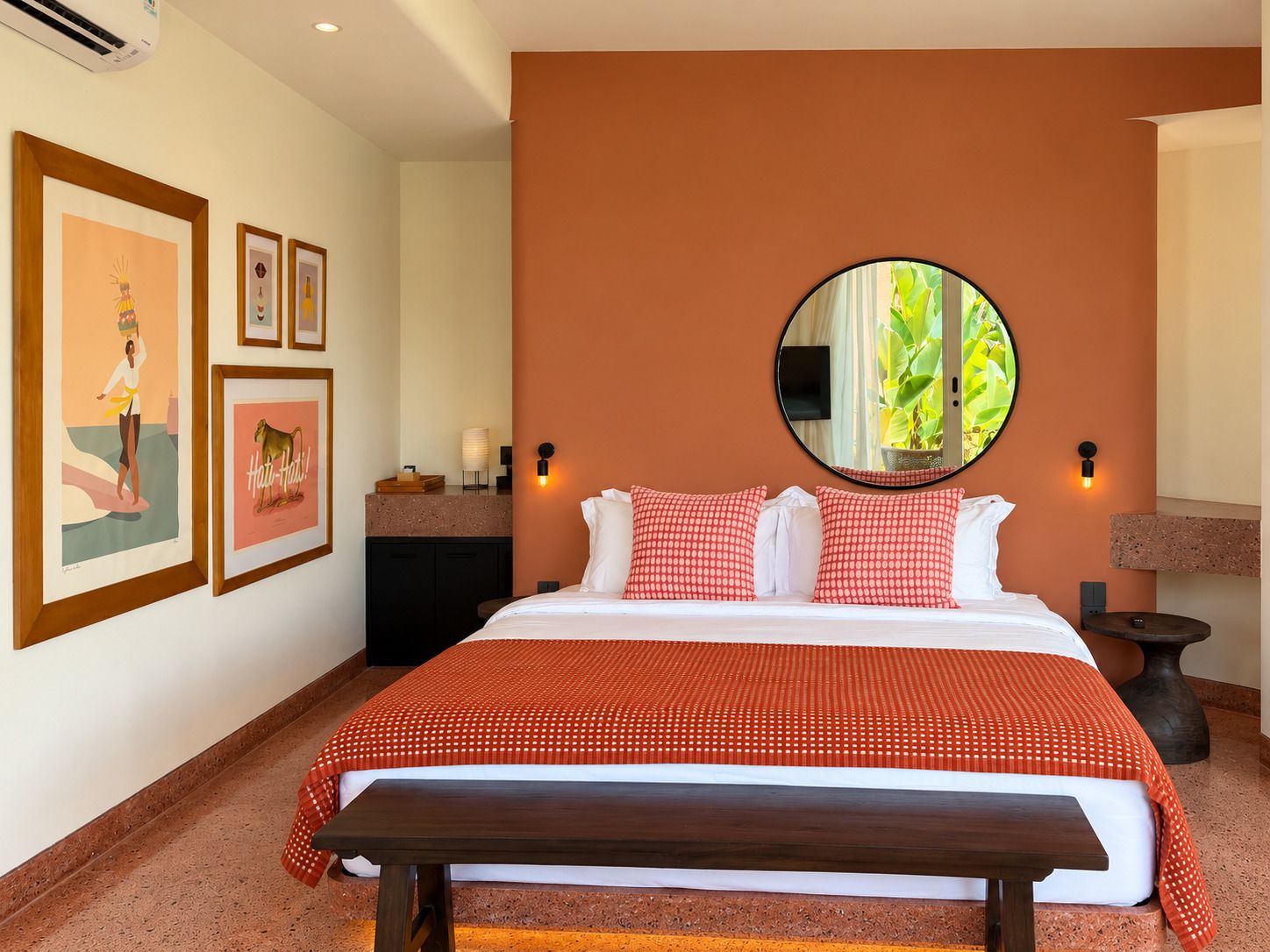 A hotel room with a bed, a round mirror, a bench, and artwork on the wall.
