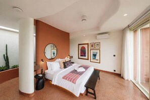 A hotel room with a bed, bedside table, bench, wall-mounted air conditioning unit, and a large window. - Villa Merah Merah Ubud by Fays Hospitality (Kabupaten Gianyar)