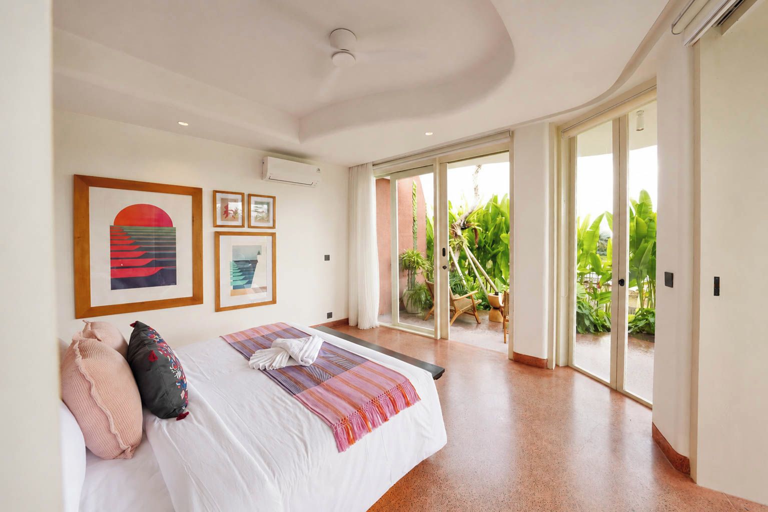 Un dormitorio con una cama grande, un ventanal corredizo que da a un balcón con plantas y cuadros decorativos en la pared.
