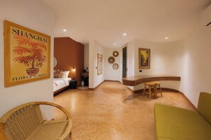 A hotel room with a bed, a sitting area with a chair, a small table, and a curved counter. There are wall decorations and a poster on the wall. - Villa Merah Merah Ubud by Fays Hospitality (Kabupaten Gianyar)