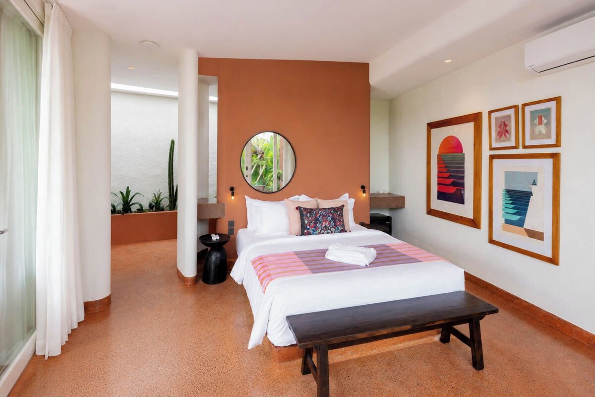 Quarto de hotel com cama, mesas de cabeceira, espelho redondo, obras de arte na parede e um banco.