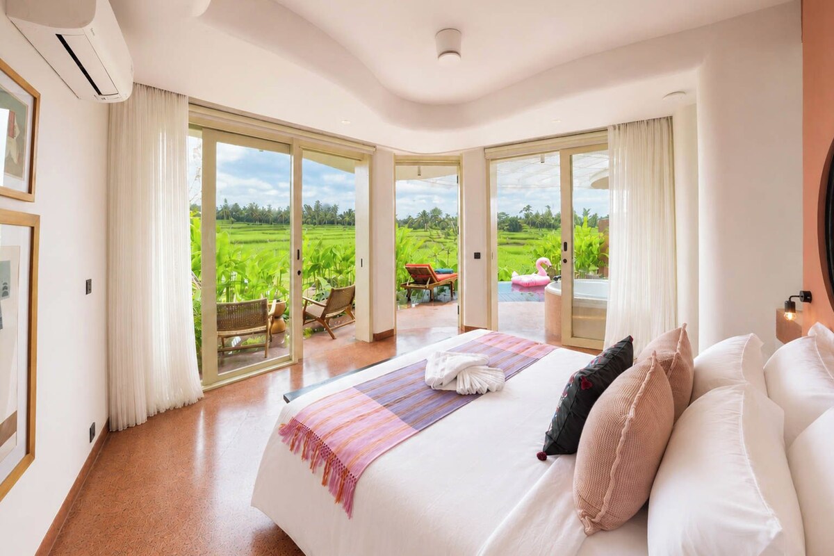 Quarto de hotel com uma cama grande, vista para uma paisagem verdejante através de portas de vidro deslizantes e varanda com mobiliário de exterior.