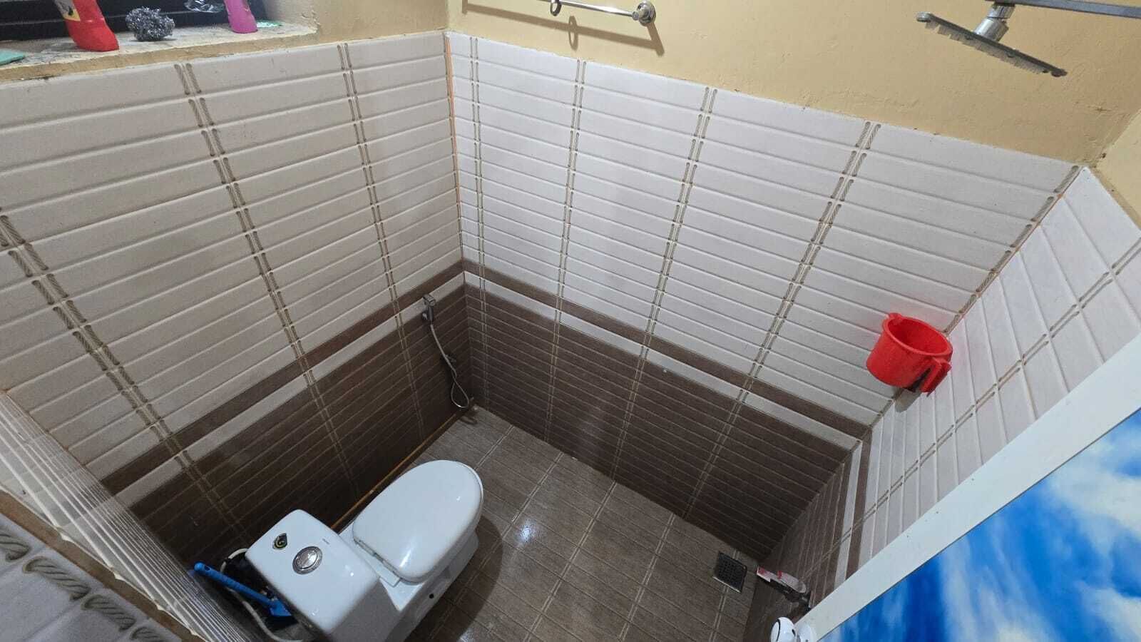 Une petite salle de bain avec des toilettes, une douche et un seau rouge accroché au mur.