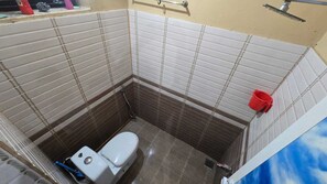A small bathroom with a toilet, a shower, and a red bucket on the wall.
