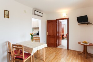 Ein Essbereich mit Tisch, Stühlen, einem kleinen Tisch mit Bananen, einem Fernseher auf einem Schrank und einer durchgehenden Tür erkennbaren Küche.