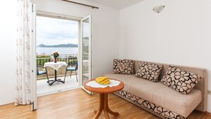 A room with a sofa, a small table with bananas, a view of the sea, and a balcony with chairs and a table. - Apartments Ana Lopud - One-bedroom Apartment With Balcony and Sea View - 4 (Dubrovnik)