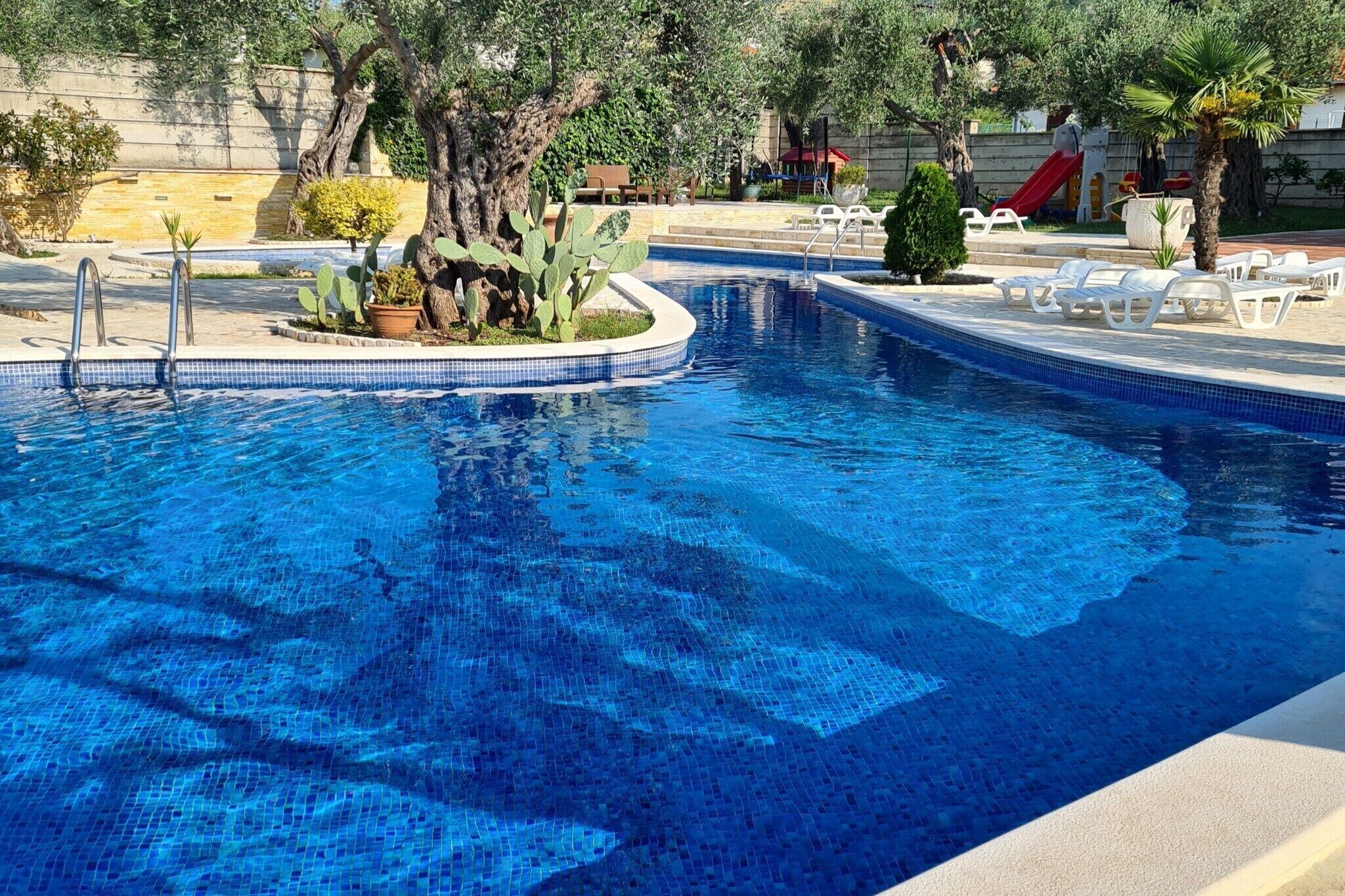 Ein Schwimmbecken mit gefliestem Rand, umgeben von Liegestühlen und Kübelpflanzen.