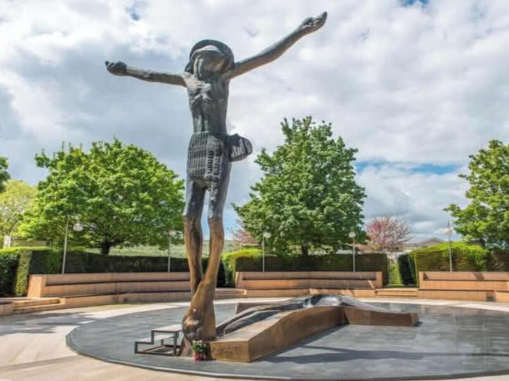 Une sculpture représentant une figure aux bras tendus, placée dans un espace public extérieur, agrémenté d’arbres et de bancs.