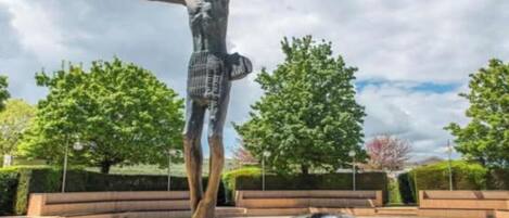 Une sculpture représentant une figure aux bras tendus, placée dans un espace public extérieur, agrémenté d’arbres et de bancs.