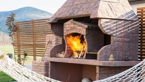 A stone outdoor pizza oven with a fire burning inside, surrounded by a wooden fence and a hammock. - Glory Sanctuary Retreat - Family room (Medugorje)