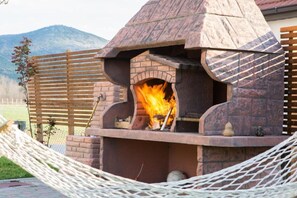 A stone outdoor pizza oven with a fire burning inside, surrounded by a wooden fence and a hammock. - Glory Sanctuary Retreat - Family room (Medugorje)