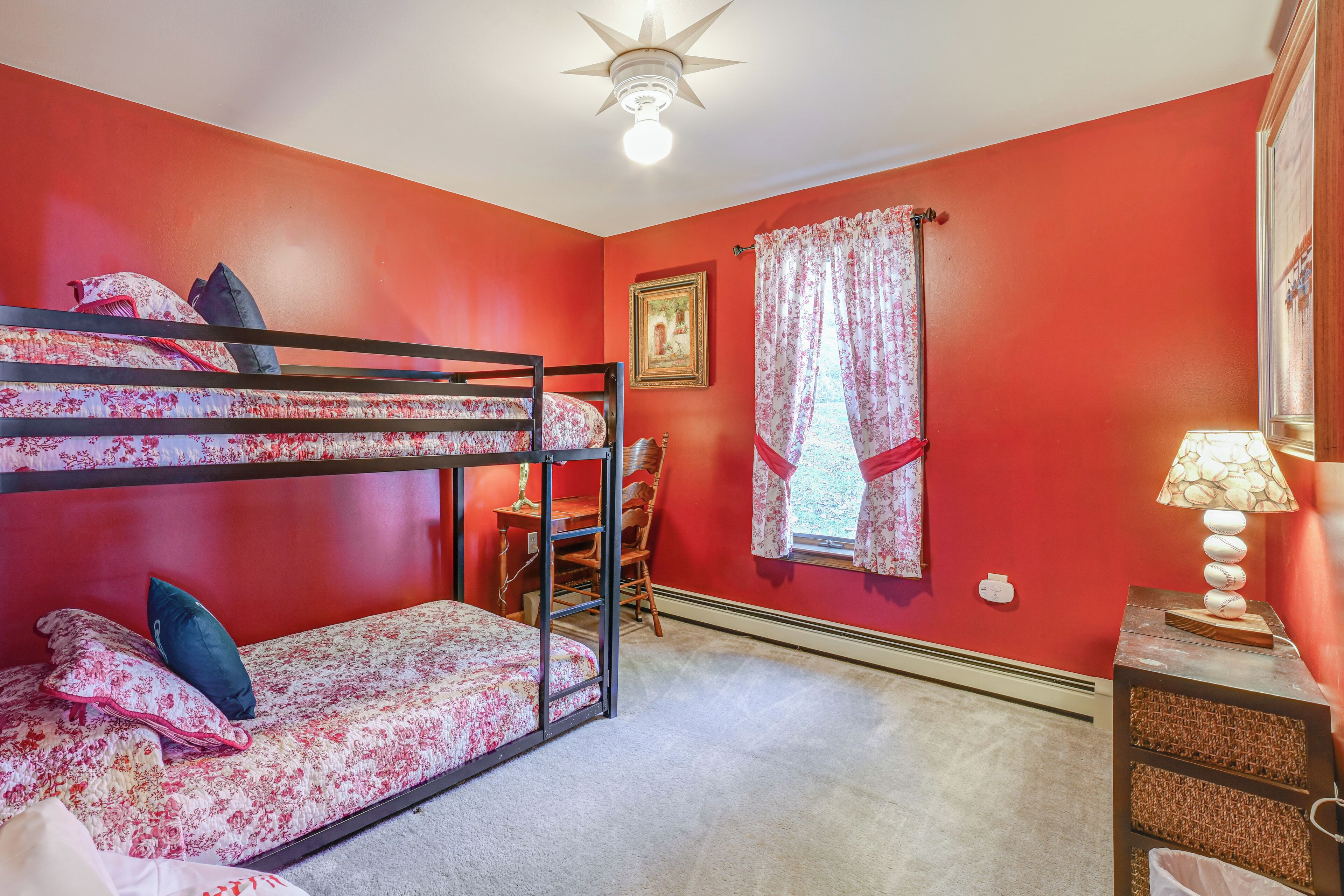 Une pièce aux murs rouges, avec un lit superposé, un bureau, une chaise, une lampe et un ventilateur de plafond.
