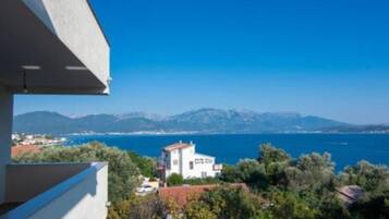 A coastal view with a mountain range in the background, a white building with a red roof, and lush greenery.