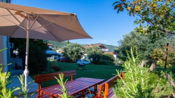 Un patio con mesa y sillas de madera, un gran paraguas y vistas a un jardín y casas al fondo.