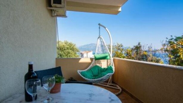 A balcony with a hanging chair, a table with a bottle and glasses, and a view of the sea and mountains. - Apartments Savić near the Sea 3 (Herceg Novi)