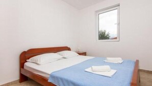 Una cama individual con cabecera de madera, ropa de cama blanca y colcha azul. Hay una mesita de noche con una lámpara y una ventana sobre la cama.