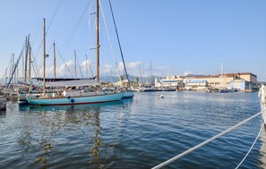 Eine Marina mit mehreren Segelbooten, die an ihren Liegeplätzen vor Anker liegen, ein Gebäude mit der Aufschrift 'AB VYCTS' im Hintergrund und ruhigem Wasser, das die Boote spiegelt.