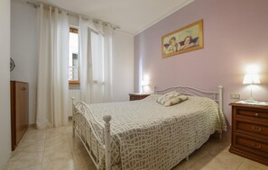 Ein Schlafzimmer mit Bett, Nachttischen, einem Schminktisch und einem Fenster mit Vorhängen, sowie einem an der Wand hängenden Bild.