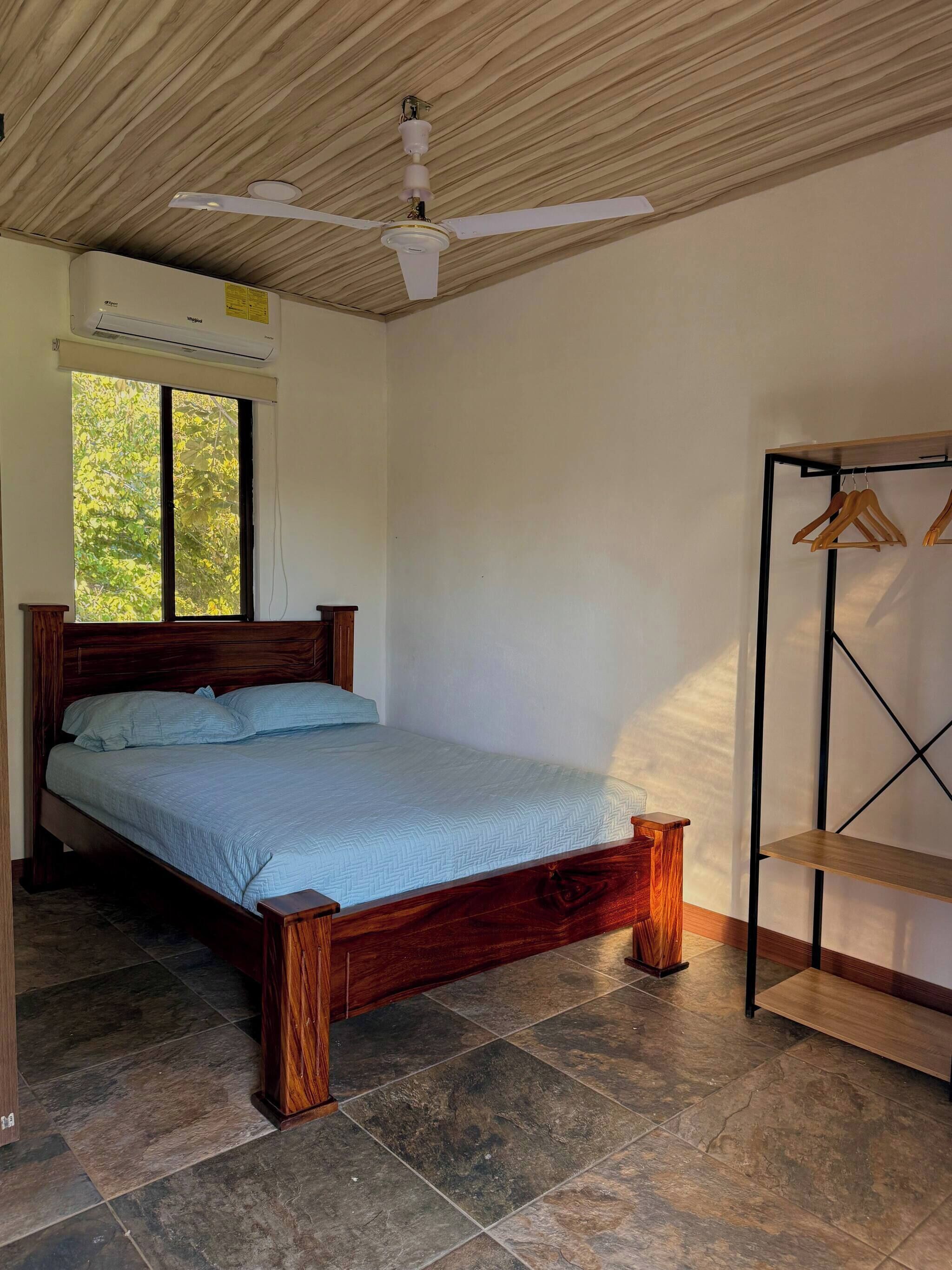 Ein Schlafzimmer mit einem hölzernen Bett, einem Deckenventilator, einer Klimaanlage und einem Fenster mit Blick auf Bäume.