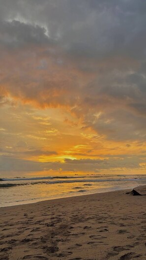 A beach with footprints leading to the water, a cloudy sky, and the sun setting or rising. - Niza 2 Bedroom house Hilltop view Saltwater Pool (Lajas de Matapalo)
