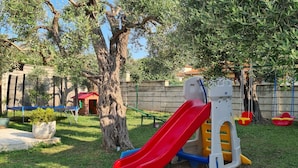 Ein Spielplatz mit einer roten Rutsche, einer Kletterkonstruktion und einer Blockhütte im Hintergrund.