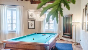 Une table de billard dans une pièce au plancher en bois, une fenêtre avec des rideaux et un tableau encadré au mur.