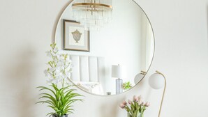 Une console blanche, surmontée d’un miroir rond, agrémentée d’une plante en pot, d’un vase de fleurs et d’une lampe.
