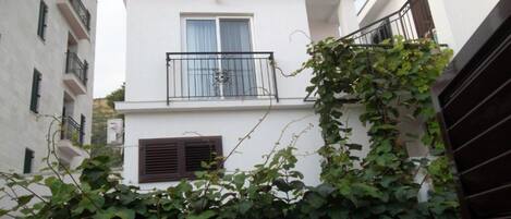 A white building with a balcony, a window with curtains, and a door with a vent.