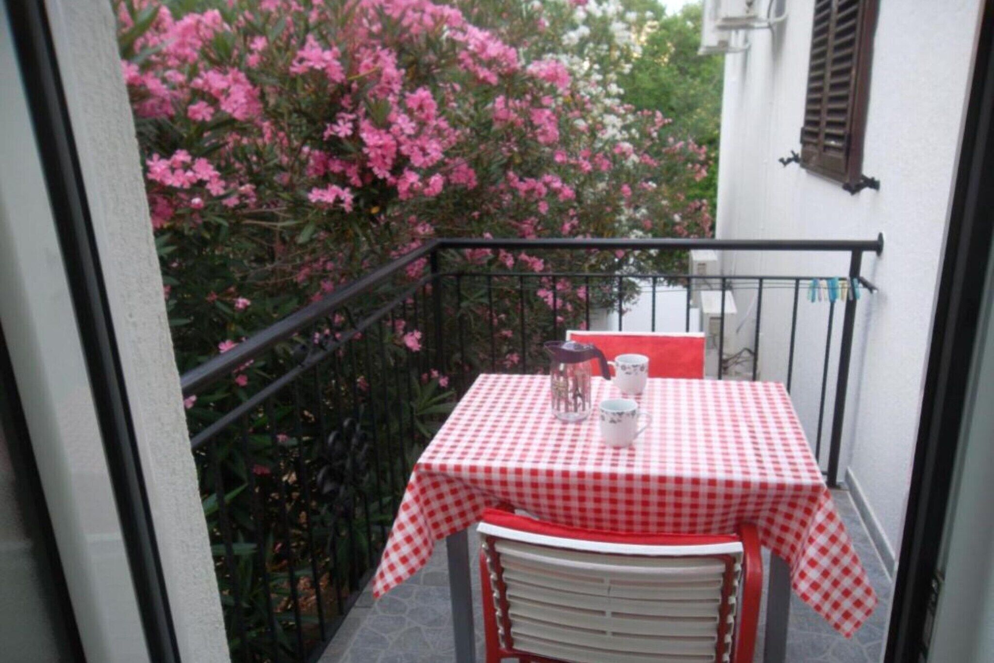 En lille balkon med et ternet bord og stole, en potteplante og et vindue med skodder.