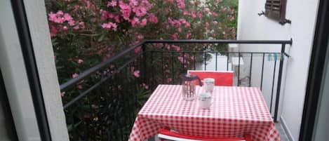 A small balcony with a checkered table and chairs, a potted plant, and a window with shutters.