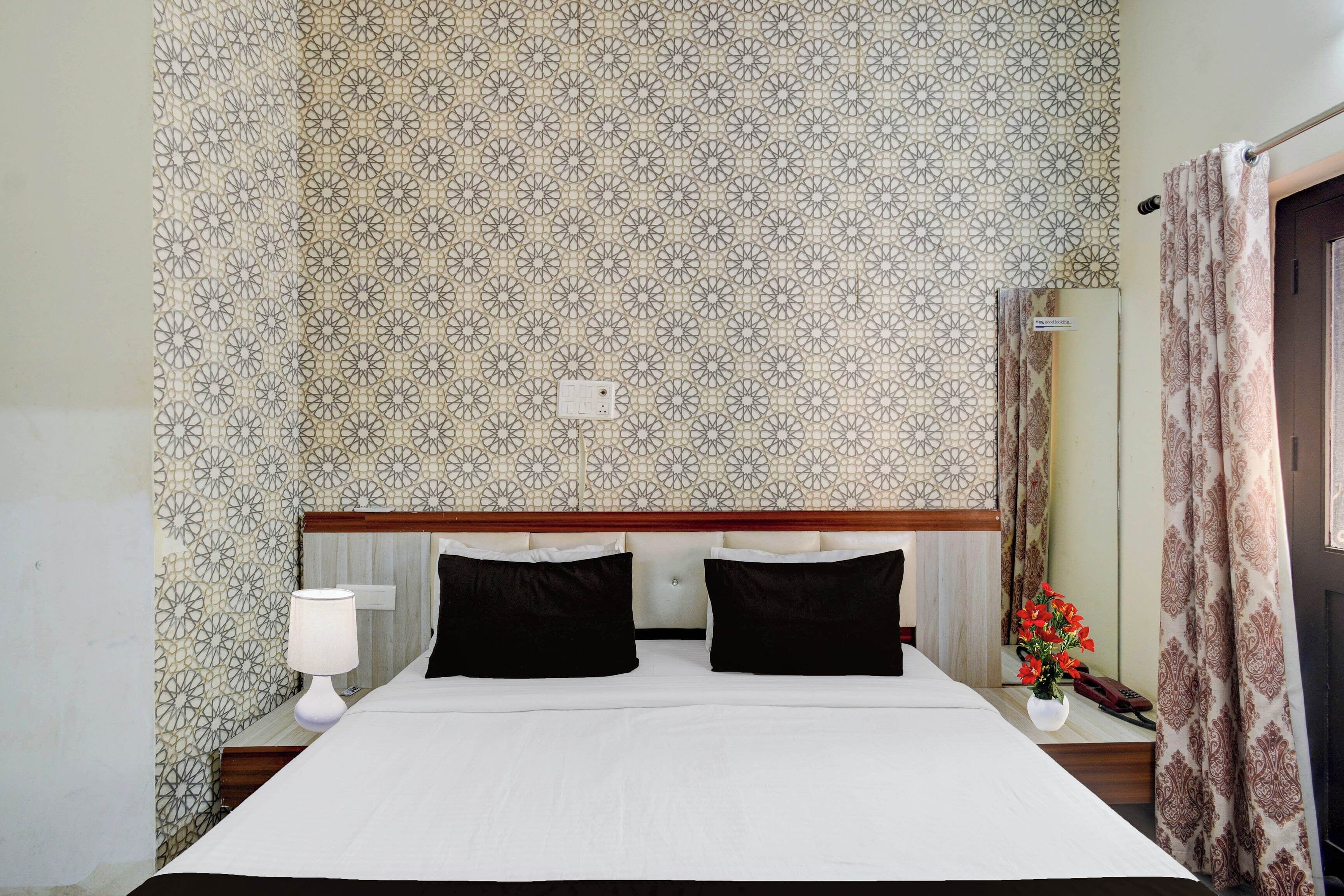 A hotel room with a patterned wallpaper, a bed with black and white bedding, a bedside table with a lamp, and a vase with flowers.