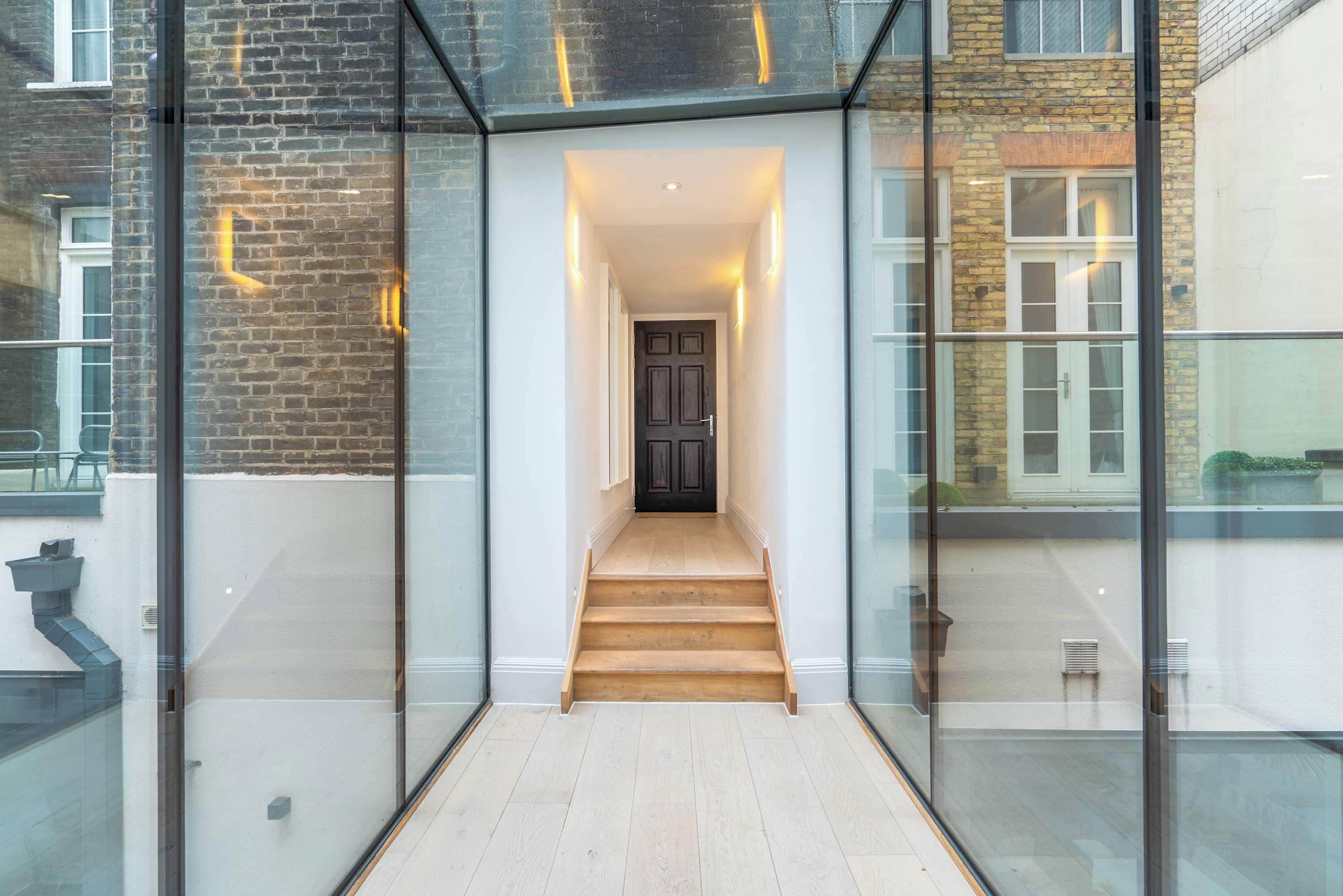 Un intérieur moderne avec un couloir vitré menant à un escalier en bois et à une porte sombre.