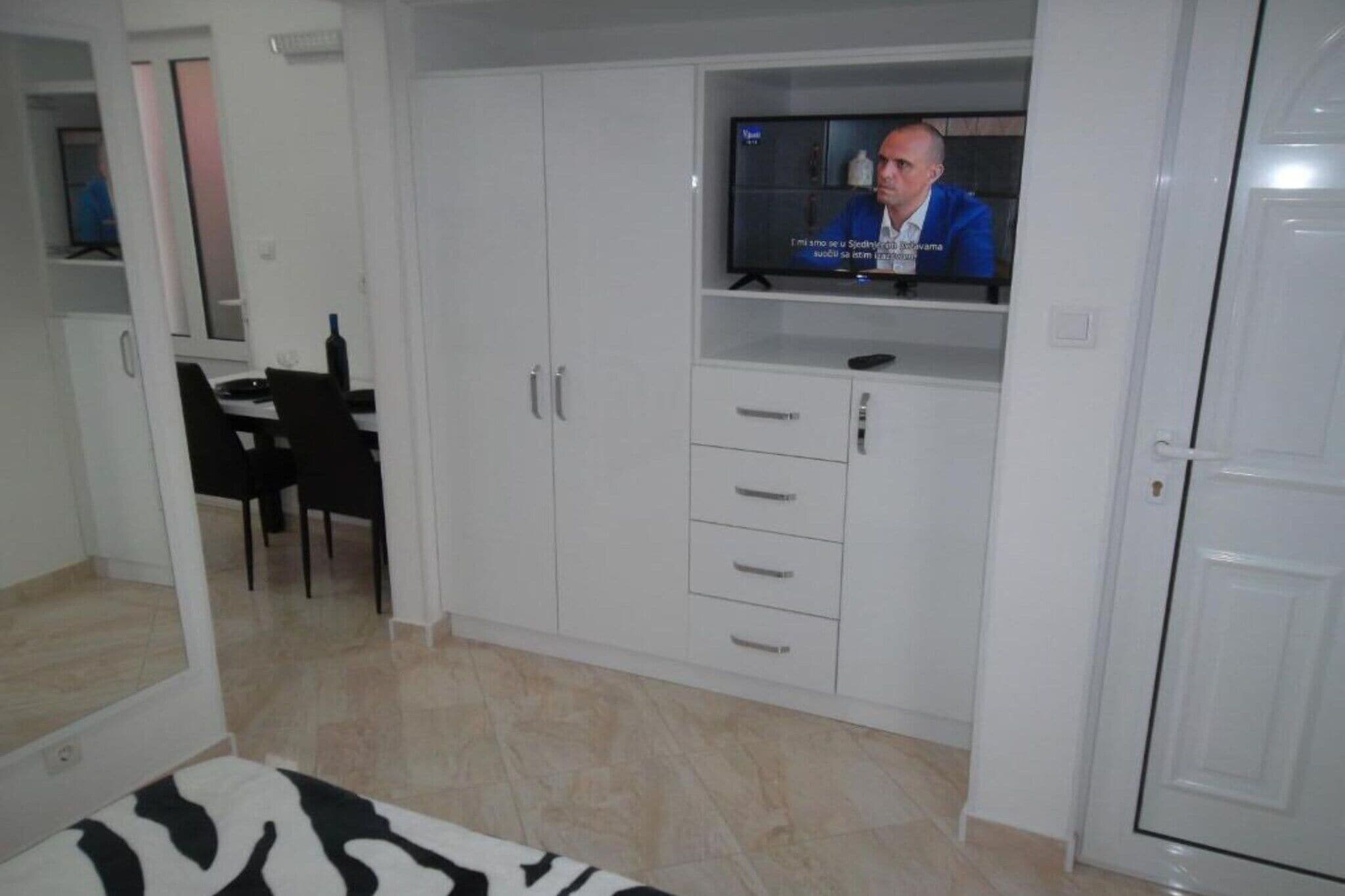 A room with a television on a stand, a dining table with chairs, and a wardrobe with multiple drawers.