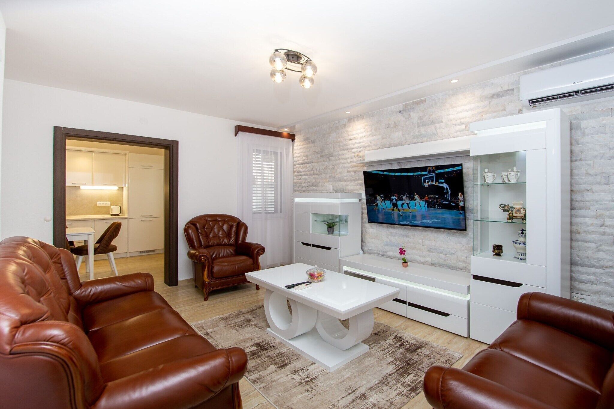 Un soggiorno moderno con divani in pelle, un tavolino da caffè bianco, una TV a schermo piatto montata su una parete con effetto pietra e una cucina visibile attraverso una porta aperta.