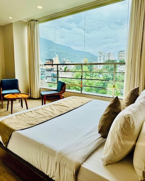 A hotel room with a large bed, two chairs, and a view of a cityscape through a large window. - The Royalistiq (Rishikesh)