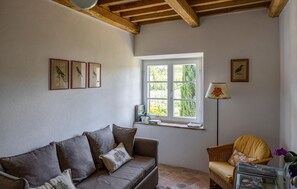 Ein gemütliches Wohnzimmer mit einer Couch, einem Sessel, einem Fenster mit Blick auf Grünflächen und gerahmten Bildern an der Wand.