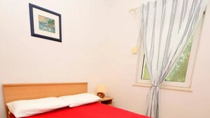 A single bed with a red bedspread, a nightstand with a lamp, a framed picture on the wall, and a window with curtains. - Coastal Breeze House - House (Selca)