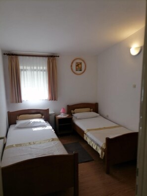 A small, simple room with two single beds, a nightstand with a lamp, a window with curtains, and a wall-mounted picture. - Apartments Libertas - Two Bedroom Apartment With Balcony and Sea View - (Rogoznica)