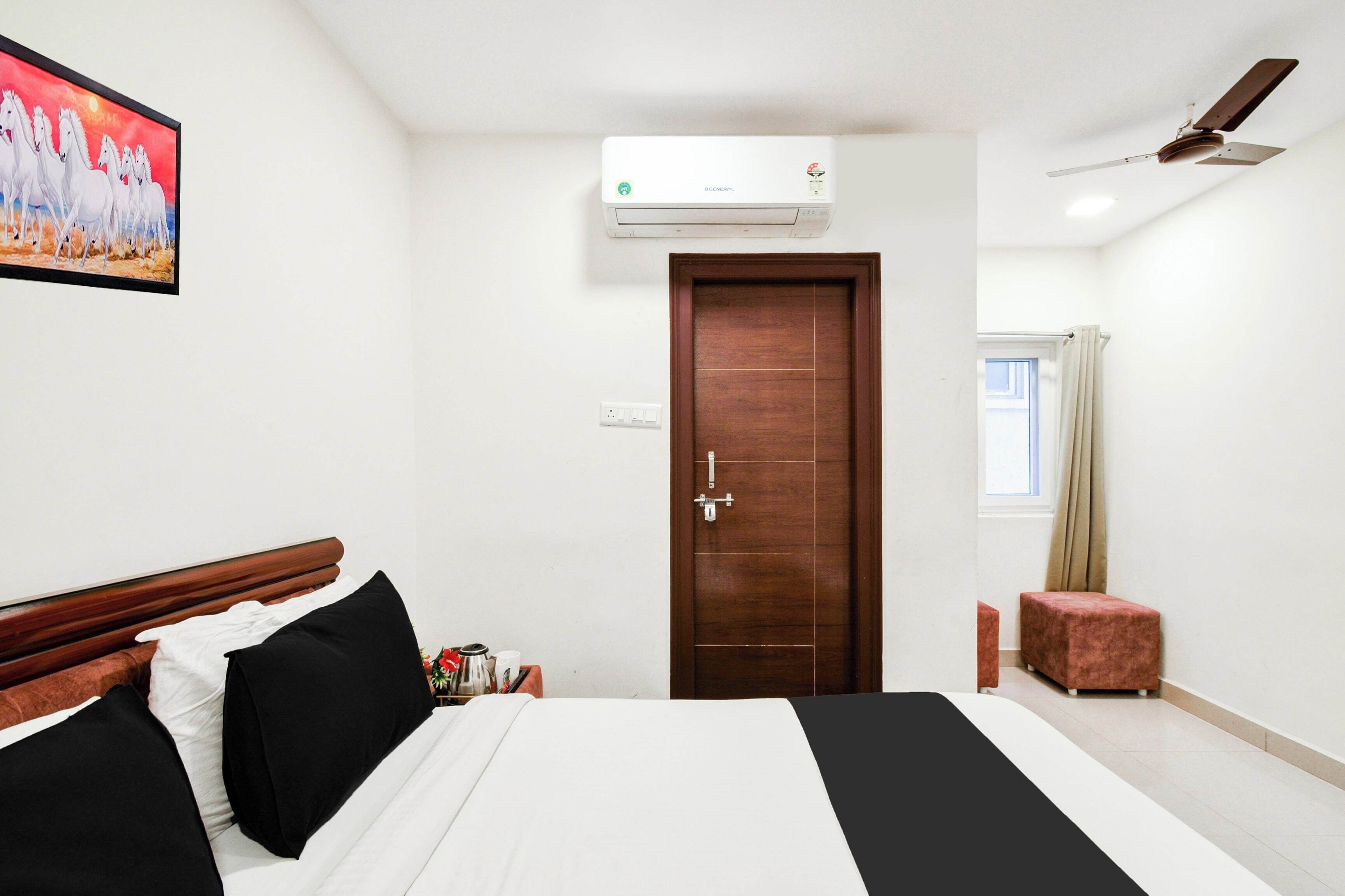 Une chambre d’hôtel avec un lit, une porte en bois, un climatiseur, un ventilateur de plafond et un tableau au mur.